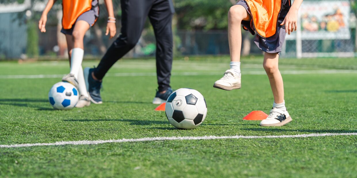 A ball on the grass between training cones, a child in an orange bib beside it.