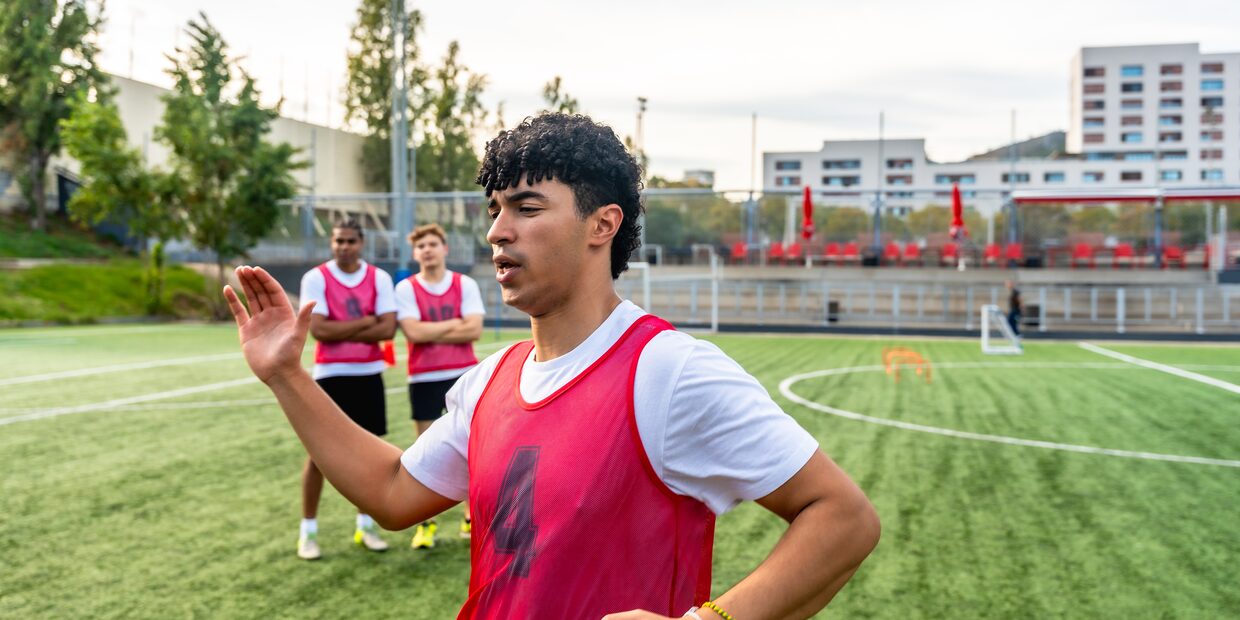 A young player in a red bib mid-sentence on the pitch, teammates standing behind him.