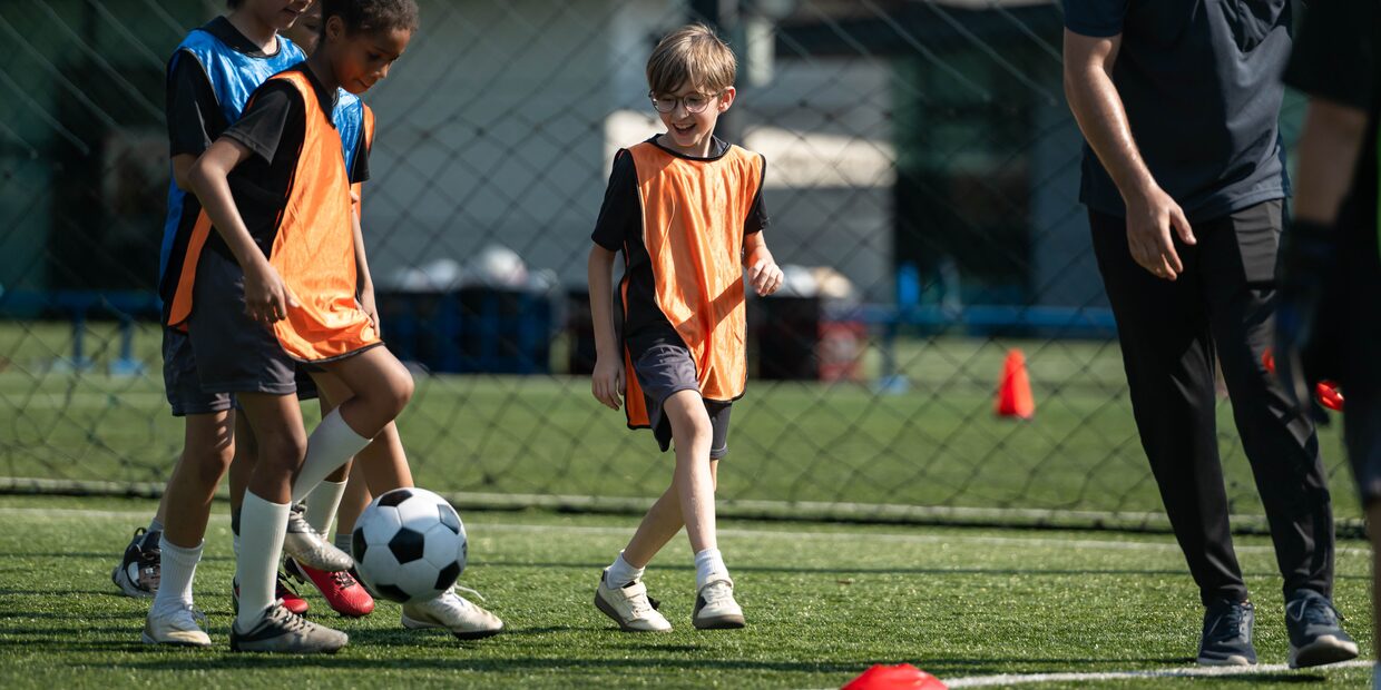 A coach on a pitch with a group of school-age children, one of them holding a ball.