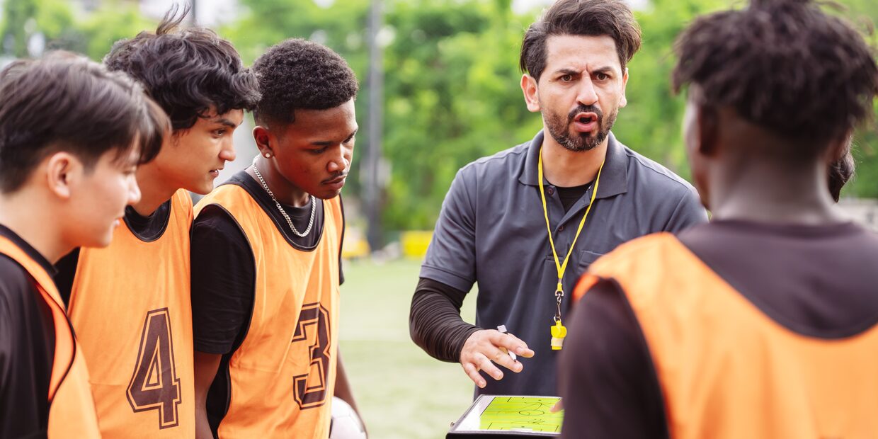 A coach with a whistle talking to three young players in numbered bibs, a clipboard in front of him.
