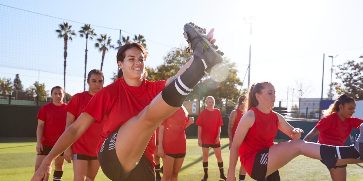 A women’s team stretching on a sunlit pitch, palm trees behind them, in low evening light.