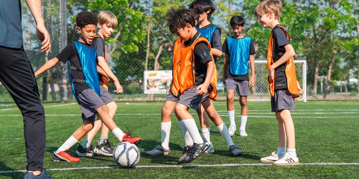 A line of children in orange and blue training bibs standing together on a pitch, one of them holding a ball, with the low sun behind them.