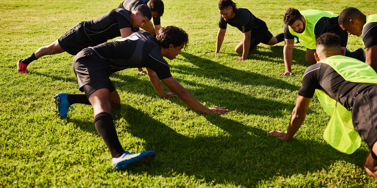 Adult players in black kit, some in yellow bibs, stretching forward in a line on the grass with long shadows across the pitch.