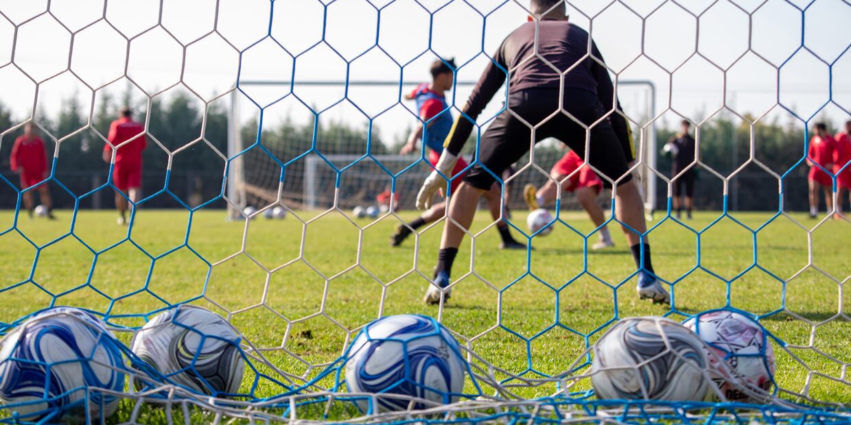 The view from inside a goal: balls resting in the net, a goalkeeper facing the play beyond it.