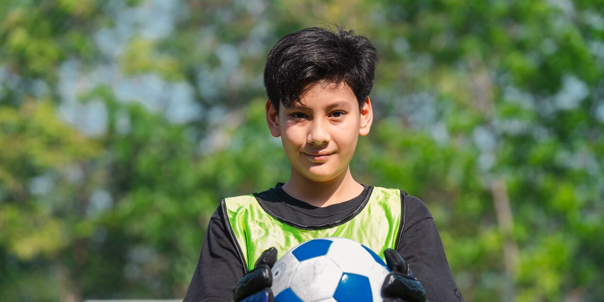 A boy in goalkeeper gloves holding a ball against his chest.