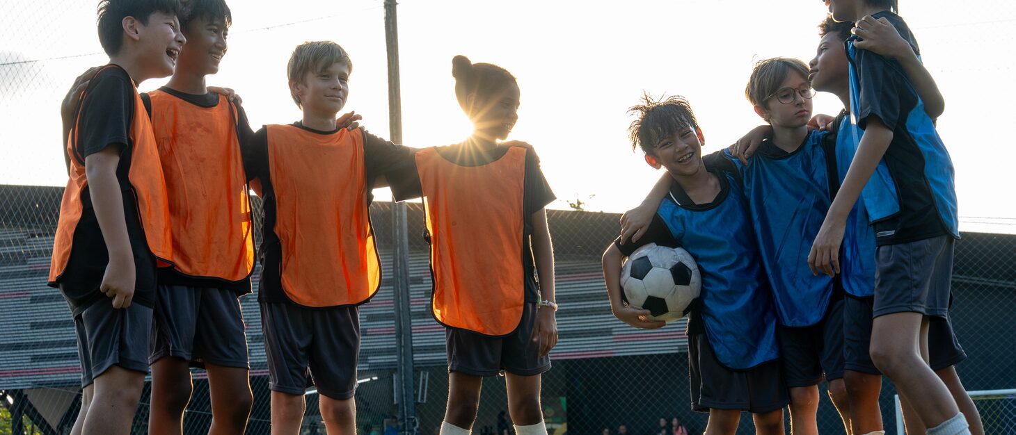 Two groups of children in orange and blue training bibs standing arm in arm on a pitch at sunset, one of them holding a ball.