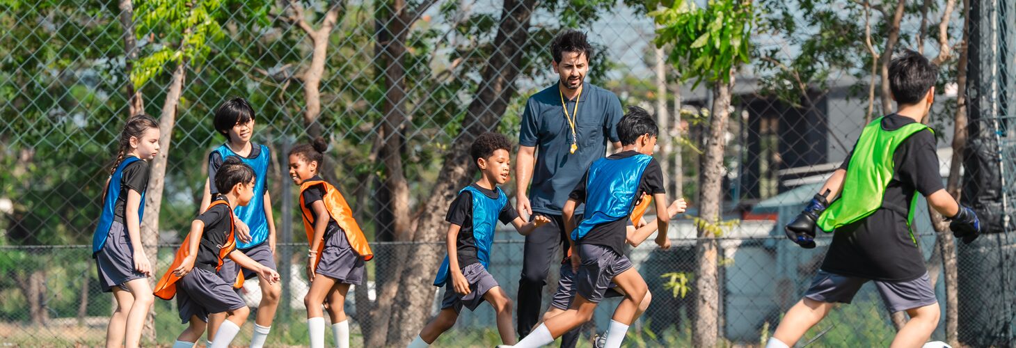 Children in blue and orange bibs playing on a pitch, a coach standing among them and trees behind.