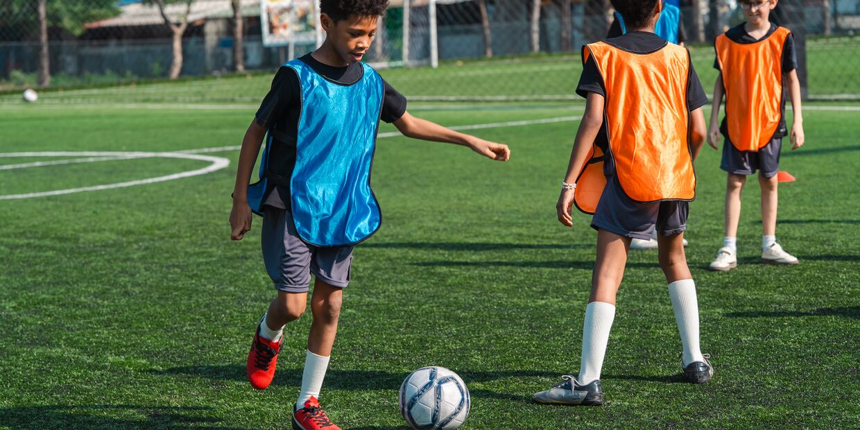 Two young players in blue and orange bibs turning with the ball on a marked pitch.